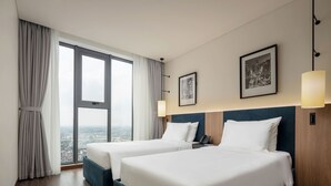 Una camera d'albergo con due letti, vista sulla città, un quadro appeso e una lampada.