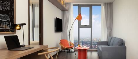 A modern living room with a wooden desk, laptop, wicker chair, grey sofa, and a view of a cityscape through large windows.