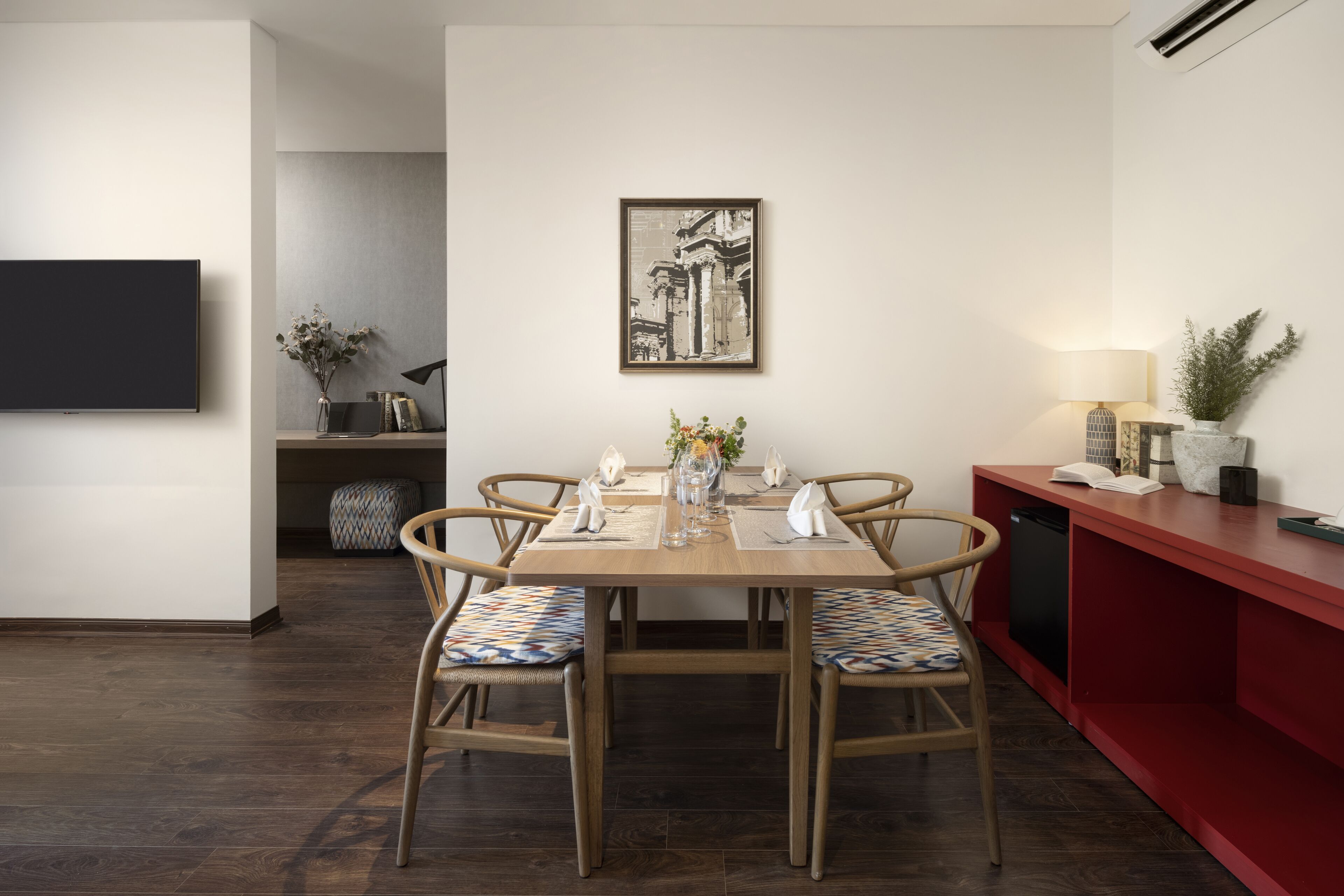 Zona pranzo con tavolo di legno apparecchiata per quattro persone, circondata da sedie in vimini con cuscini decorati. Sopra il tavolo, un quadro appeso al muro.