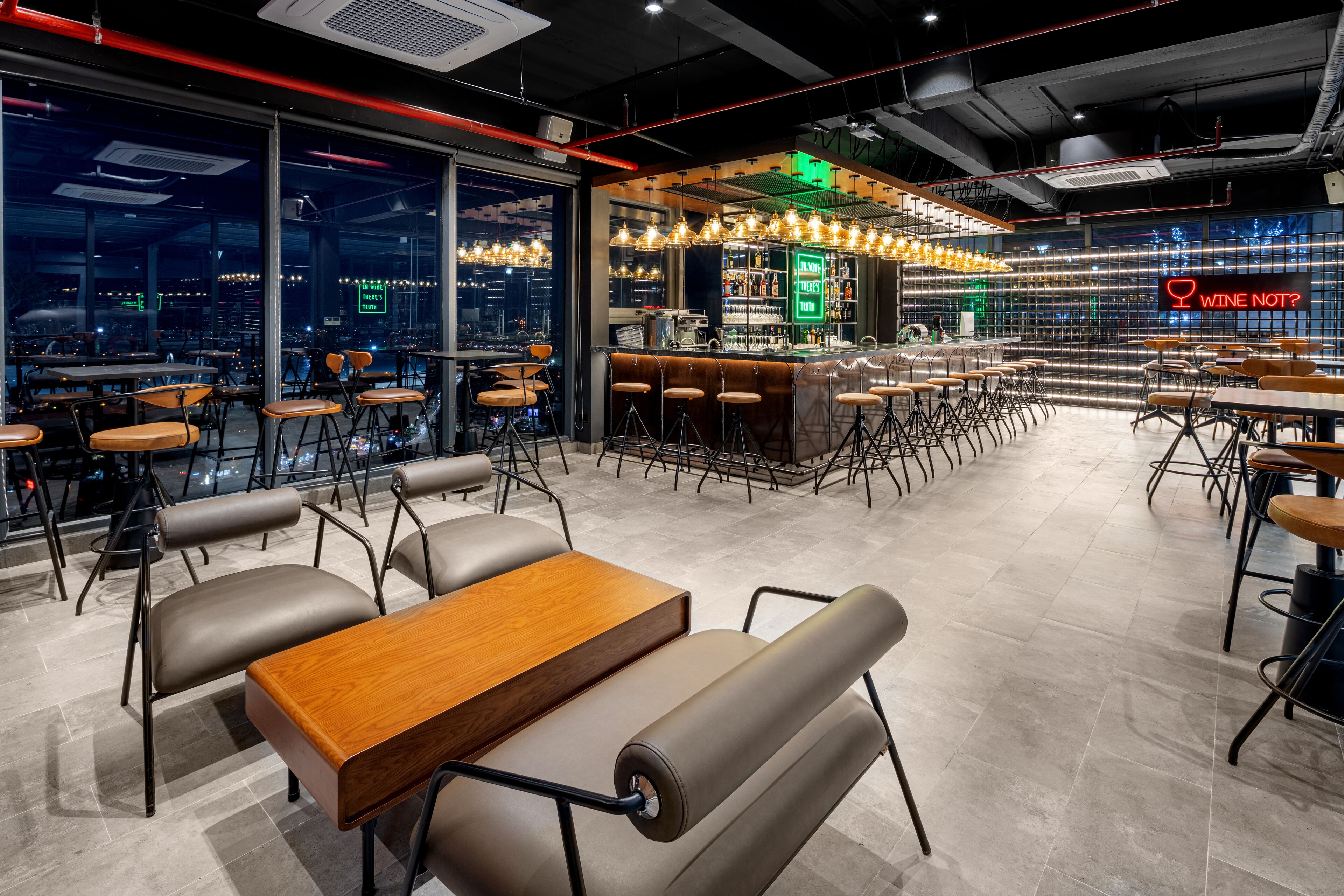 A modern bar area with a neon sign, seating arrangements, and a city view through large windows.