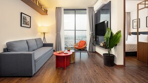 Una camera d'albergo moderna con un divano grigio, un tavolino rosso, una sedia in vimini, una grande finestra con tende, un televisore a schermo piatto e una pianta in vaso.