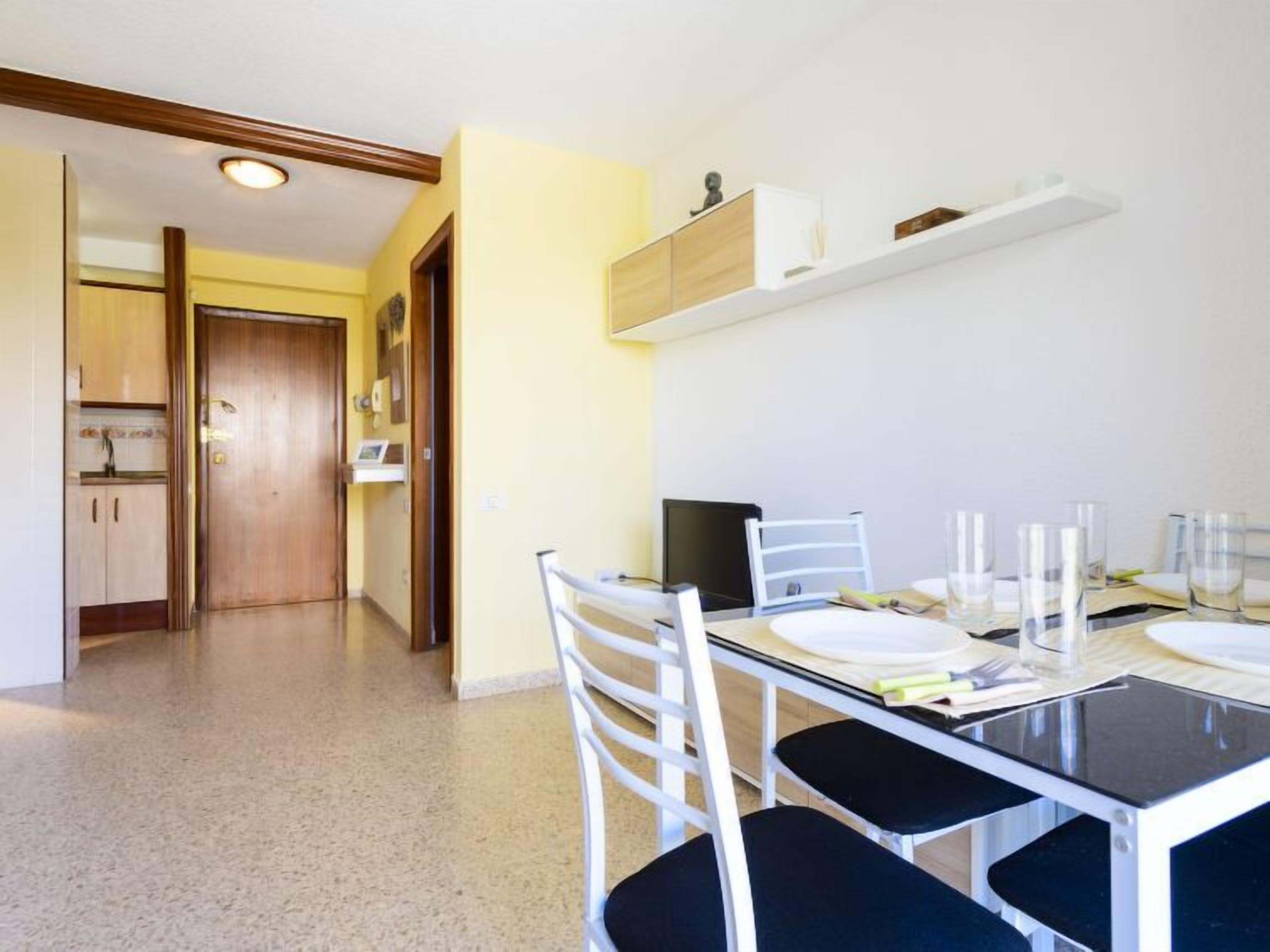 A dining area with a glass table, white chairs, and a TV. There is a wooden door and a kitchen visible in the background.
