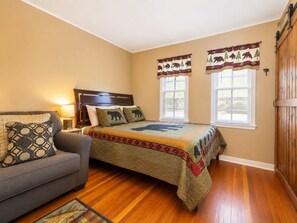 A bedroom with a bed, a sofa, two windows with curtains, and a wooden floor.