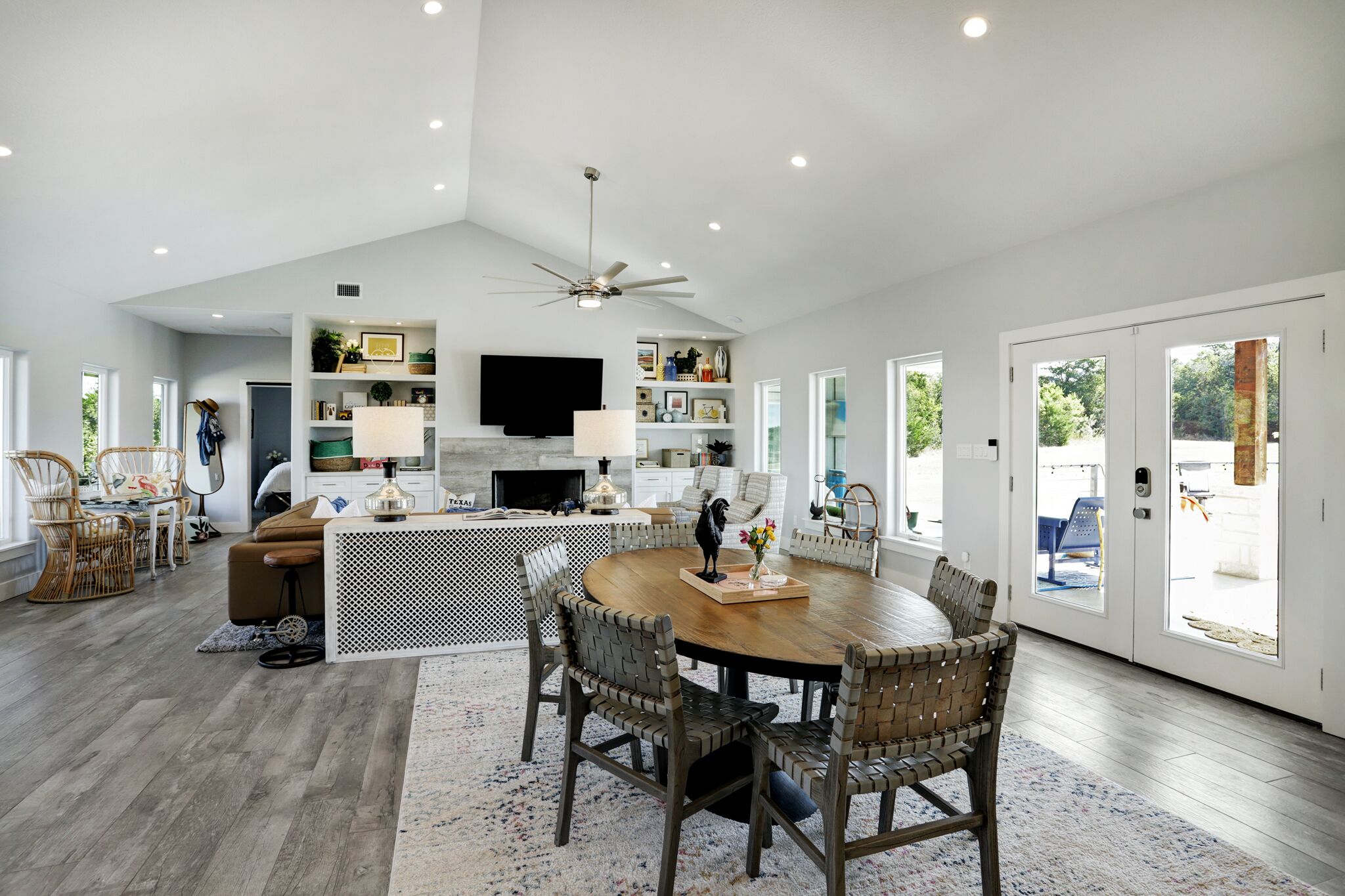 A spacious living area with a large round table, wicker chairs, a ceiling fan, and a TV mounted on the wall.