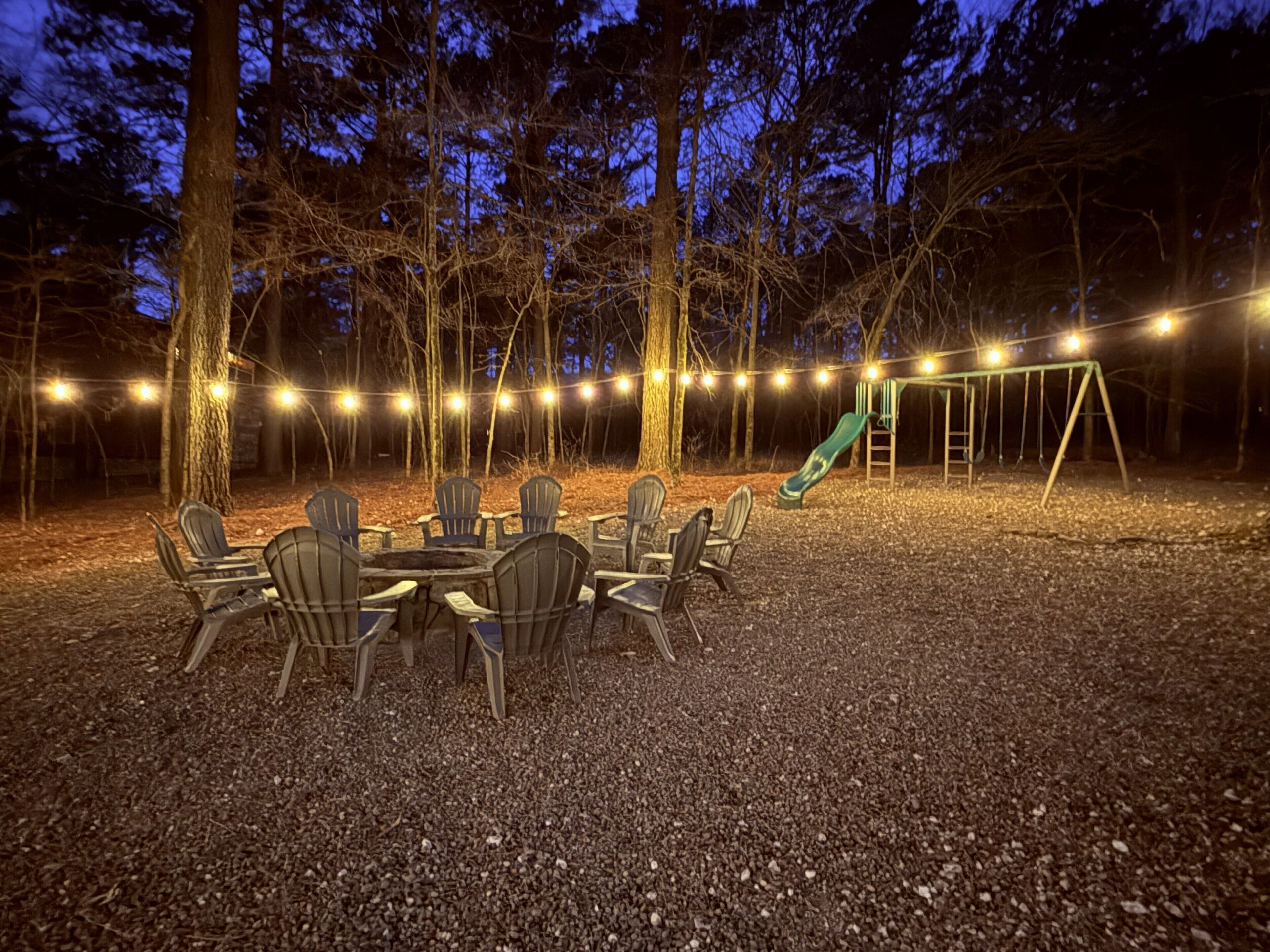 Un espace extérieur aménagé avec des chaises et une table, entouré d’arbres et d’une balançoire, le tout illuminé par des guirlandes lumineuses.