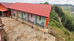 A building with a red roof and multiple rooms, situated on a hillside with a view of green hills and a winding path.