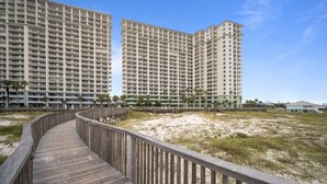 A wooden boardwalk leading to high-rise buildings with balconies, surrounded by sand and sparse vegetation. - Beach Club 1603A - Lil Bit of Paradise is your perfect beach getaway (Gulf Shores)