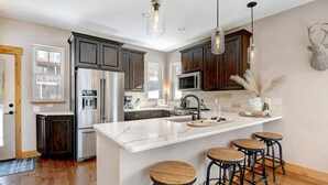 A modern kitchen with dark wood cabinets, stainless steel appliances, a white marble island with bar stools, and pendant lighting.