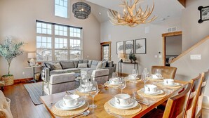A spacious living room with a large window, a dining area set for six, and a modern chandelier.