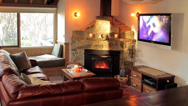 A cozy living room with a stone fireplace, leather sofa, and a television mounted on the wall. - Wallace's-Chalet (Dinner Plain)