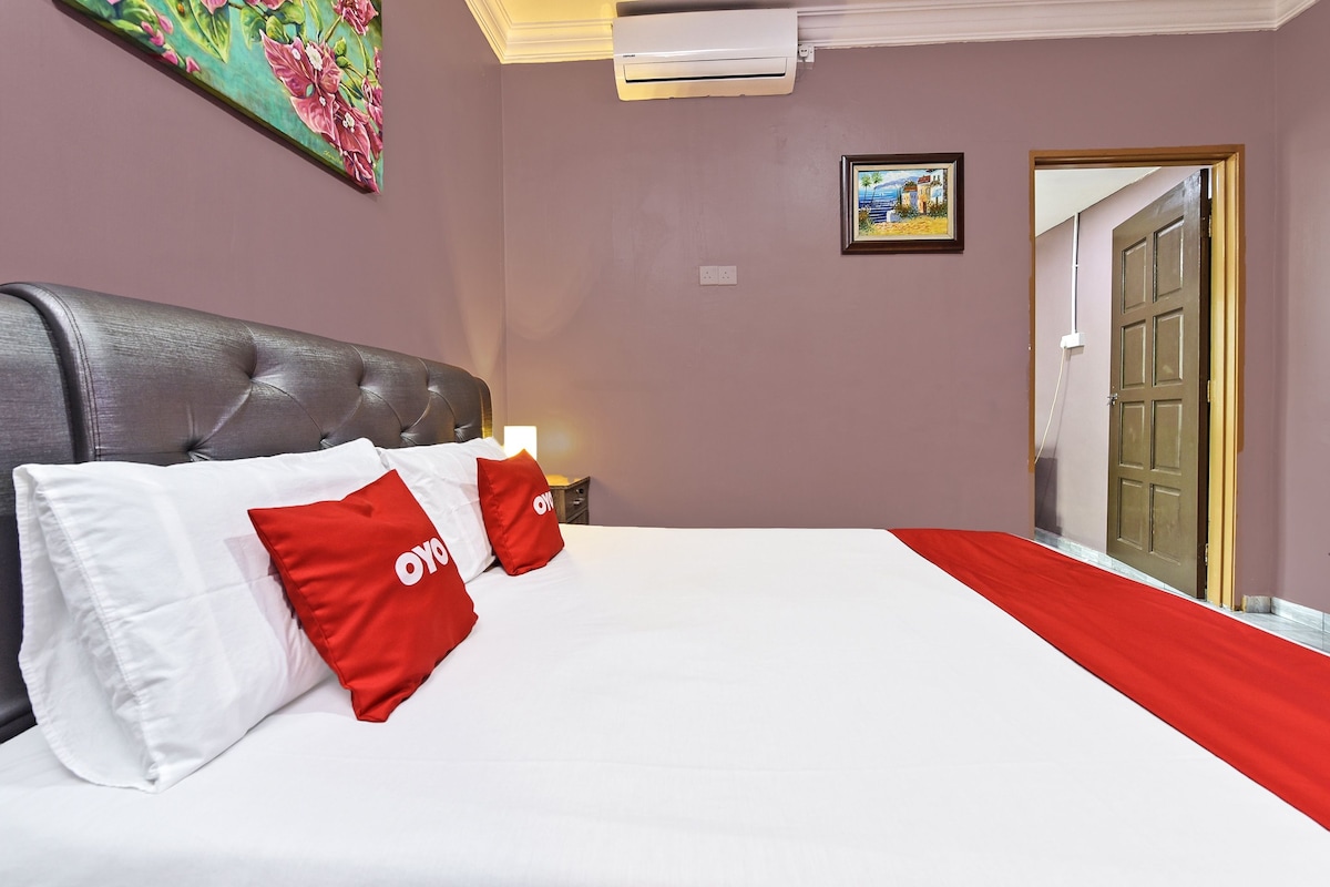 A hotel room with a large bed, a headboard, a nightstand, a lamp, a painting on the wall, and an air conditioning unit.