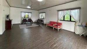 Une pièce spacieuse dotée d’un ventilateur de plafond, de grandes fenêtres avec des rideaux, d’un plancher en bois et d’un canapé agrémenté de coussins rouges.