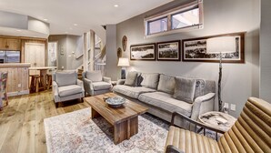 Un salon moderne comprenant un canapé, des fauteuils, une table basse et un tapis. On y trouve un escalier menant à un autre niveau, un coin cuisine avec des meubles en bois et un coin repas avec des tabourets.