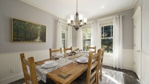 A dining room with a wooden table set for six, white chairs, a chandelier, and a painting on the wall.