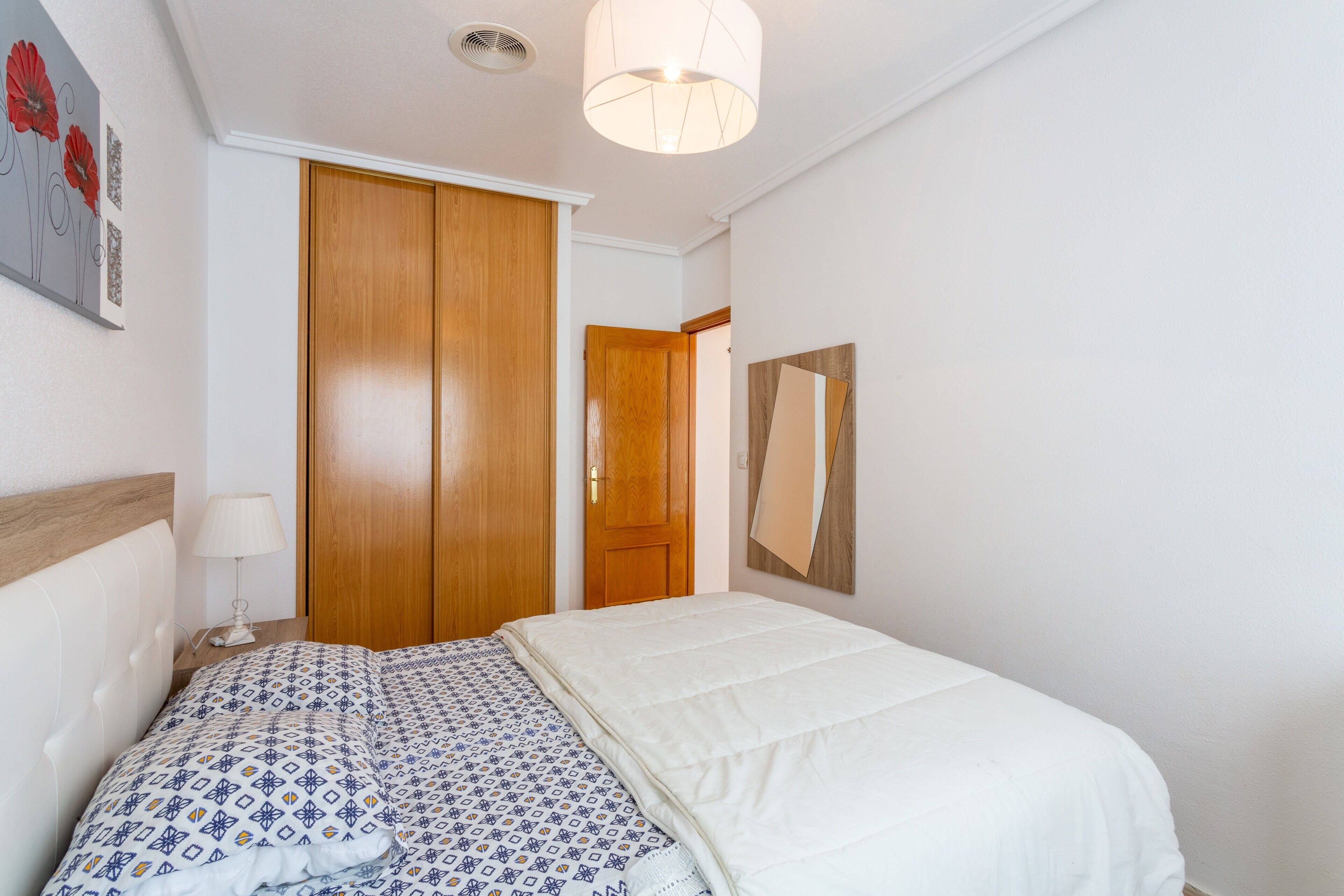 Um quarto com cama, mesa de cabeceira, luminária, guarda-roupa de madeira e luminária no teto.