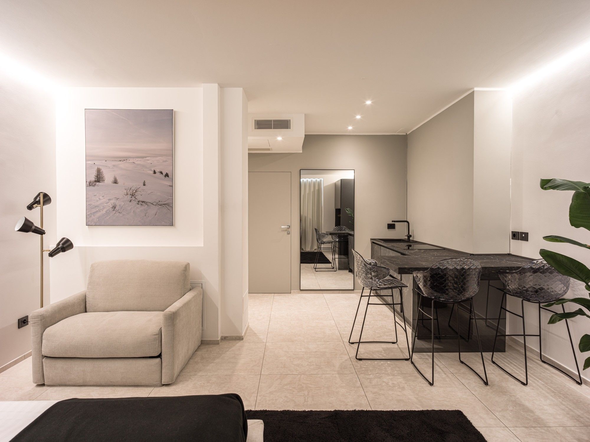 A modern living room with a sofa, a dining area with bar stools, a large mirror, and a painting of a snowy landscape.