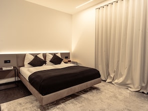 Une chambre à coucher moderne avec un lit, des tables de chevet, une petite table et une grande fenêtre avec des rideaux.