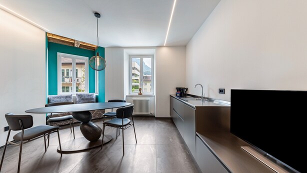 A modern kitchen with a large TV, a dining area with a round table and chairs, and a view of a building through a window.