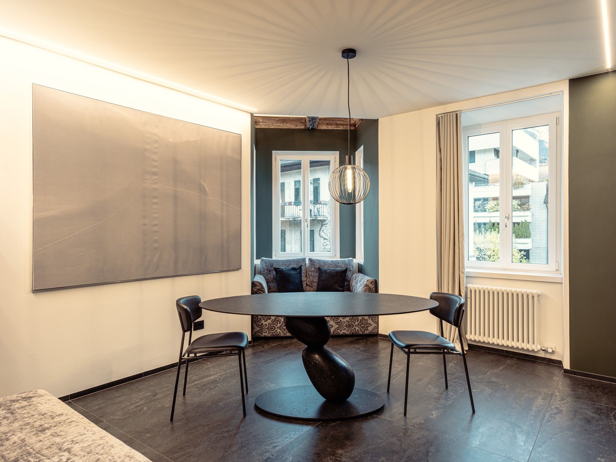 Et moderne spiseområde med et rundt bord, to stole, en hængende lampe og udsigt til bygninger gennem et vindue.