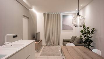 Une cuisine moderne avec un plan de travail et un évier blancs, un coin repas avec une table et des chaises en bois, un canapé avec des coussins et une grande plante.