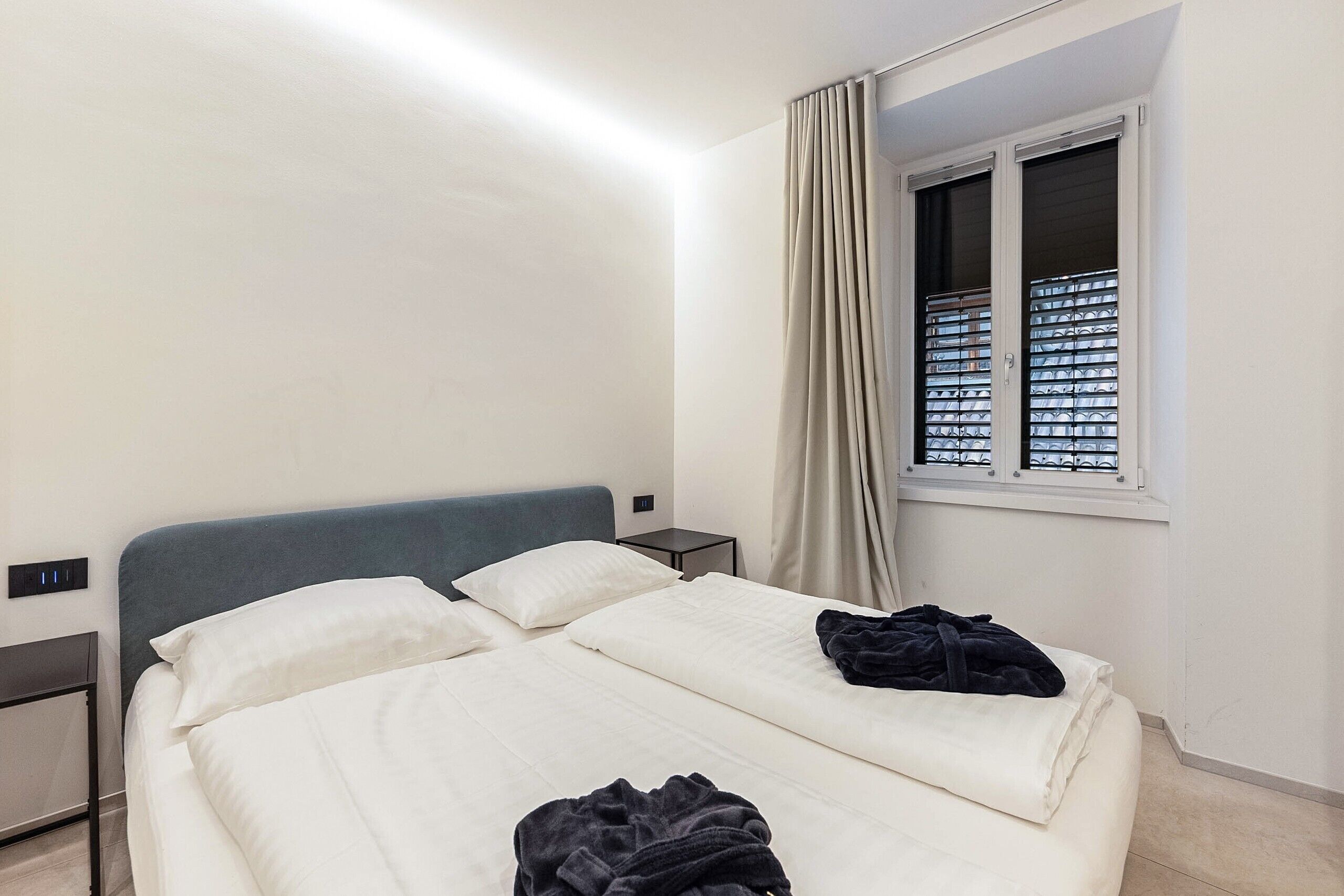 A hotel room with a bed, a bedside table, a window with blinds, and a curtain.