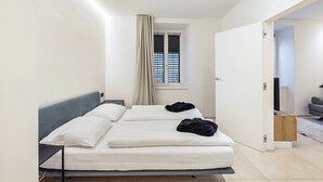 A bedroom with a bed, a window with curtains, a door leading to another room, and a small table.