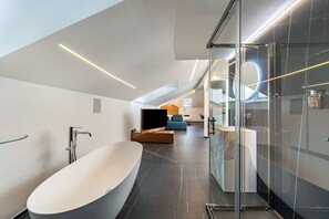 A modern bathroom with a freestanding bathtub, a glass shower enclosure, and a visible living area with a sofa and a television.