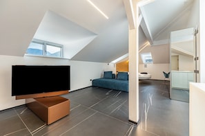 A modern loft-style room with a large flat-screen TV, a blue sofa, and a dining area with a white table and chairs.