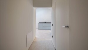 Un couloir étroit mène à une cuisine dotée de meubles blancs et d’un plan de travail noir.