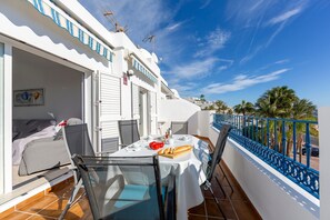 Een balkon met een eettafel, een stoel, een bed en uitzicht op palmbomen en een straat.