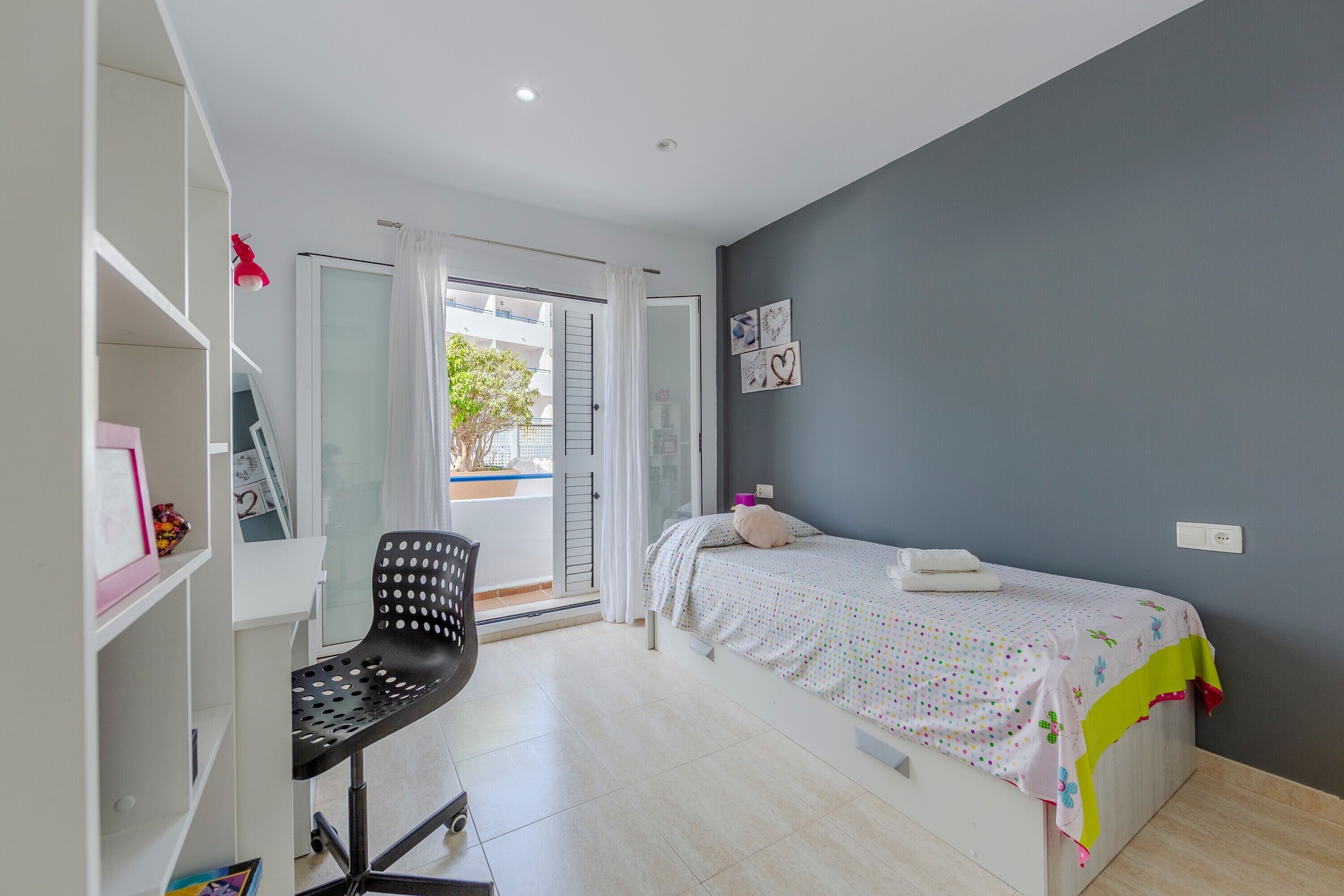 A bedroom with a bed, a desk, a chair, a wardrobe, and a view through a glass door.