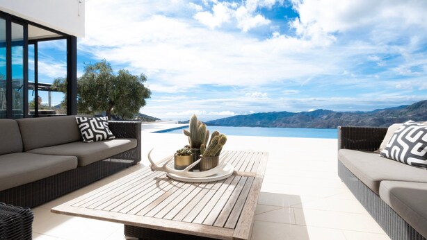 Ein moderner Außenbereich mit Holztisch, zwei Sofas und Blick auf einen See und Berge.