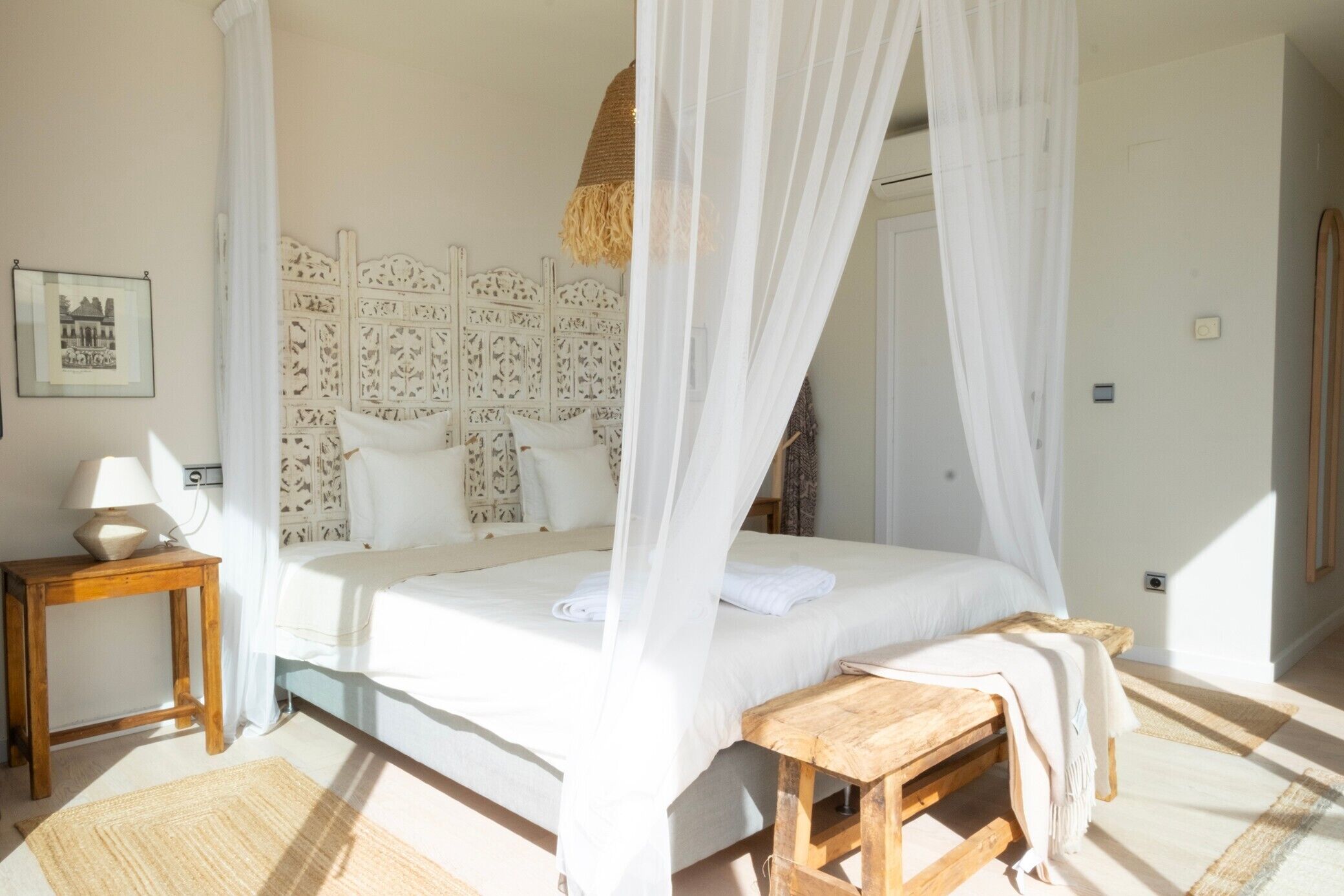 Een slaapkamer met een bed, nachtkastje, houten bankje en hemelbed.