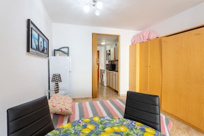 Een compacte kamer met een eettafel, twee stoelen, een gestreept vloerkleed, een kledingkast, een klein kitchenette en een deur naar een andere kamer.