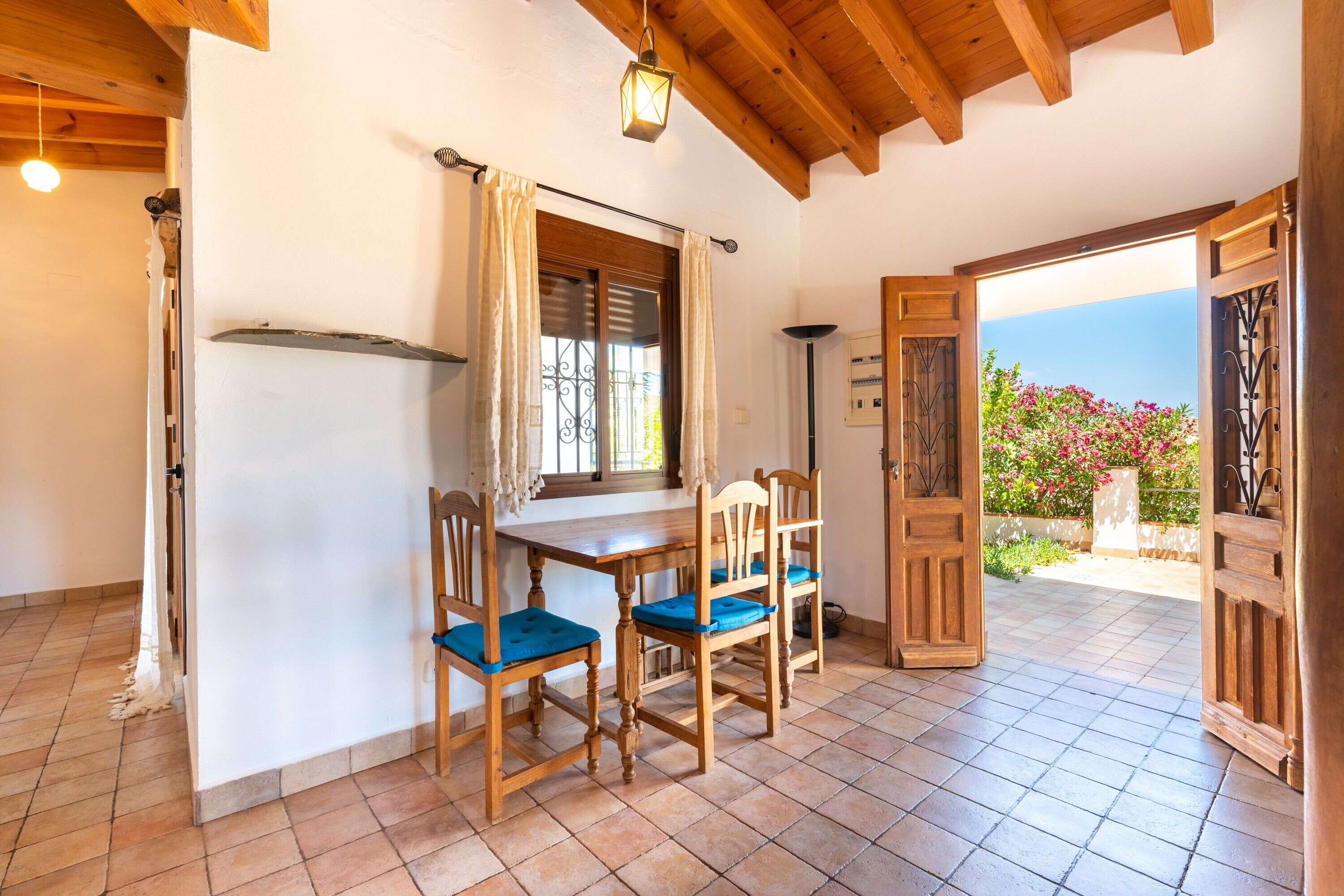 Una habitación con una mesa de madera, sillas con cojines azules, una ventana con cortinas y vistas a un jardín con flores rosadas.