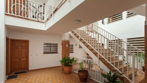 Un interior espacioso con una puerta de madera, suelo de baldosas, plantas en macetas y una escalera blanca.