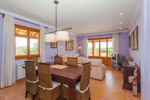 Un comedor con una mesa de madera y sillas de mimbre, un sofá y un televisor en una habitación con paredes moradas y ventanas grandes.
