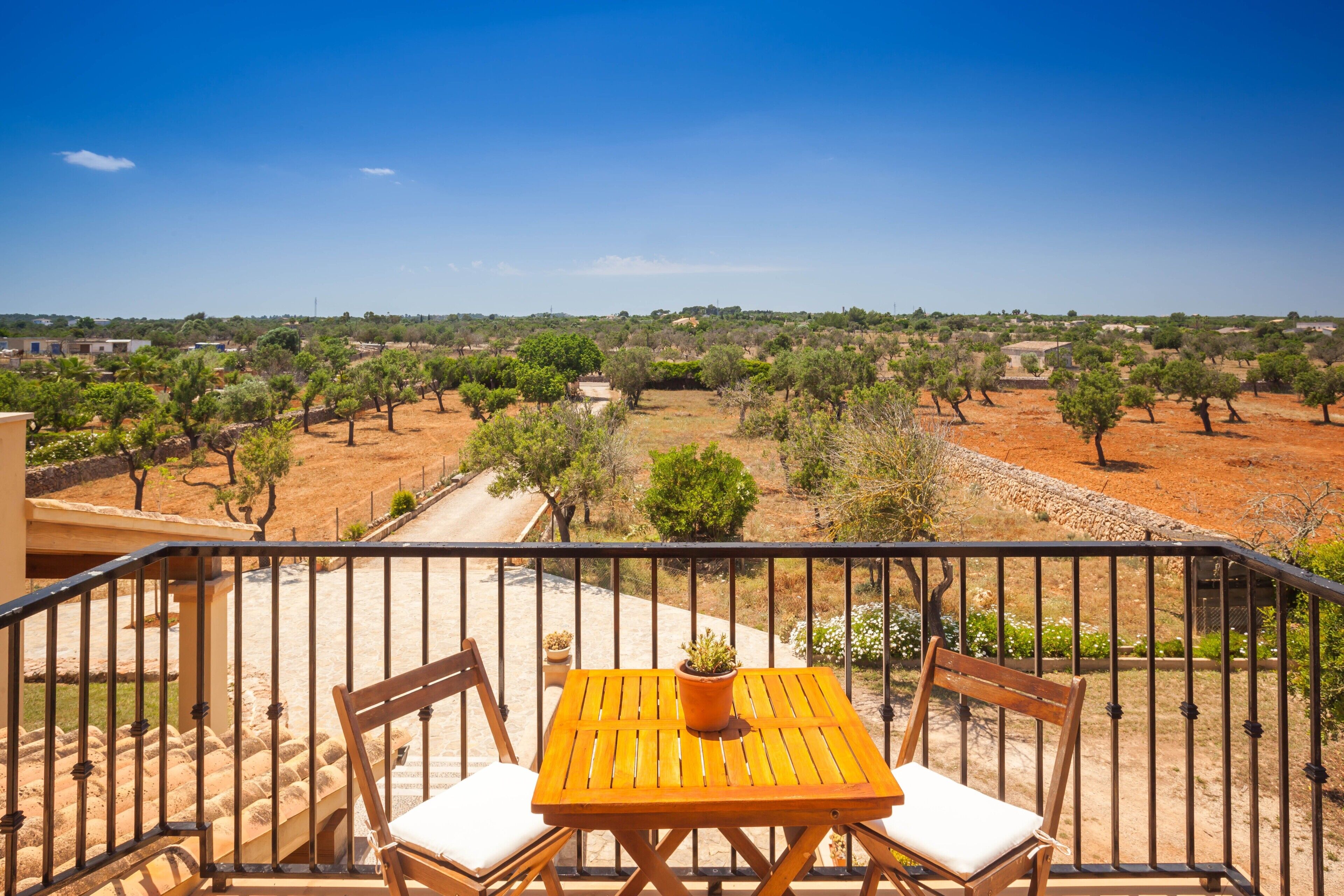 A balcony with a wooden table and chairs, overlooking a landscape with trees and a clear sky.