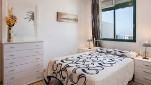 A bedroom with a bed, a dresser, a nightstand, a lamp, a window with curtains, and a cityscape painting on the wall.