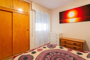 Une chambre avec une armoire en bois, un lit recouvert d’une couvre-lit à motifs, une commode blanche et un tableau représentant un coucher de soleil accroché au mur.