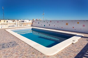 Une piscine dont le fond est recouvert de carreaux géométriques, entourée d’une terrasse dallée de pierres, et un mur blanc orné de motifs carrés colorés.
