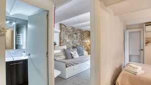A bedroom with a stone wall, a bed with a beige quilt, a white bedside table, and a bathroom with a modern sink and mirror.