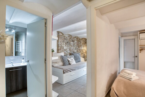 A bedroom with a stone wall, a bed with a beige quilt, a white bedside table, and a bathroom with a modern sink and mirror.