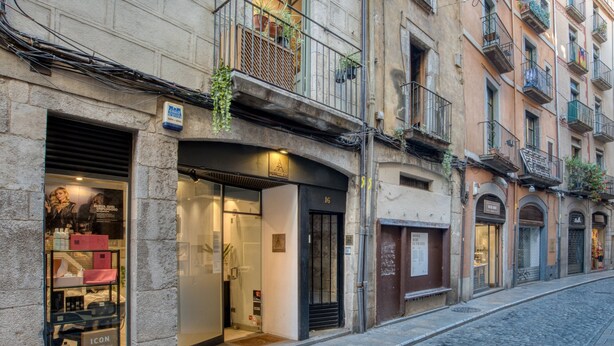 A street view with a building featuring a storefront with a glass facade, a visible entrance, and a balcony with plants.
