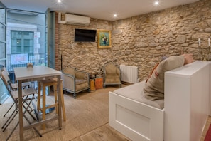 Une pièce avec un mur en pierre, un canapé blanc, une table à manger avec des chaises et un téléviseur fixé au mur.