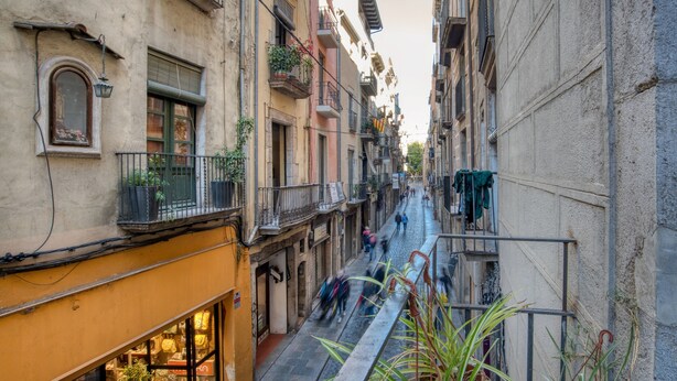 A narrow street with buildings on either side, balconies with plants, and a shop with glass doors. - Apartment “Old Side Girona one” with Balcony, Wi-Fi & A/C (Girona)