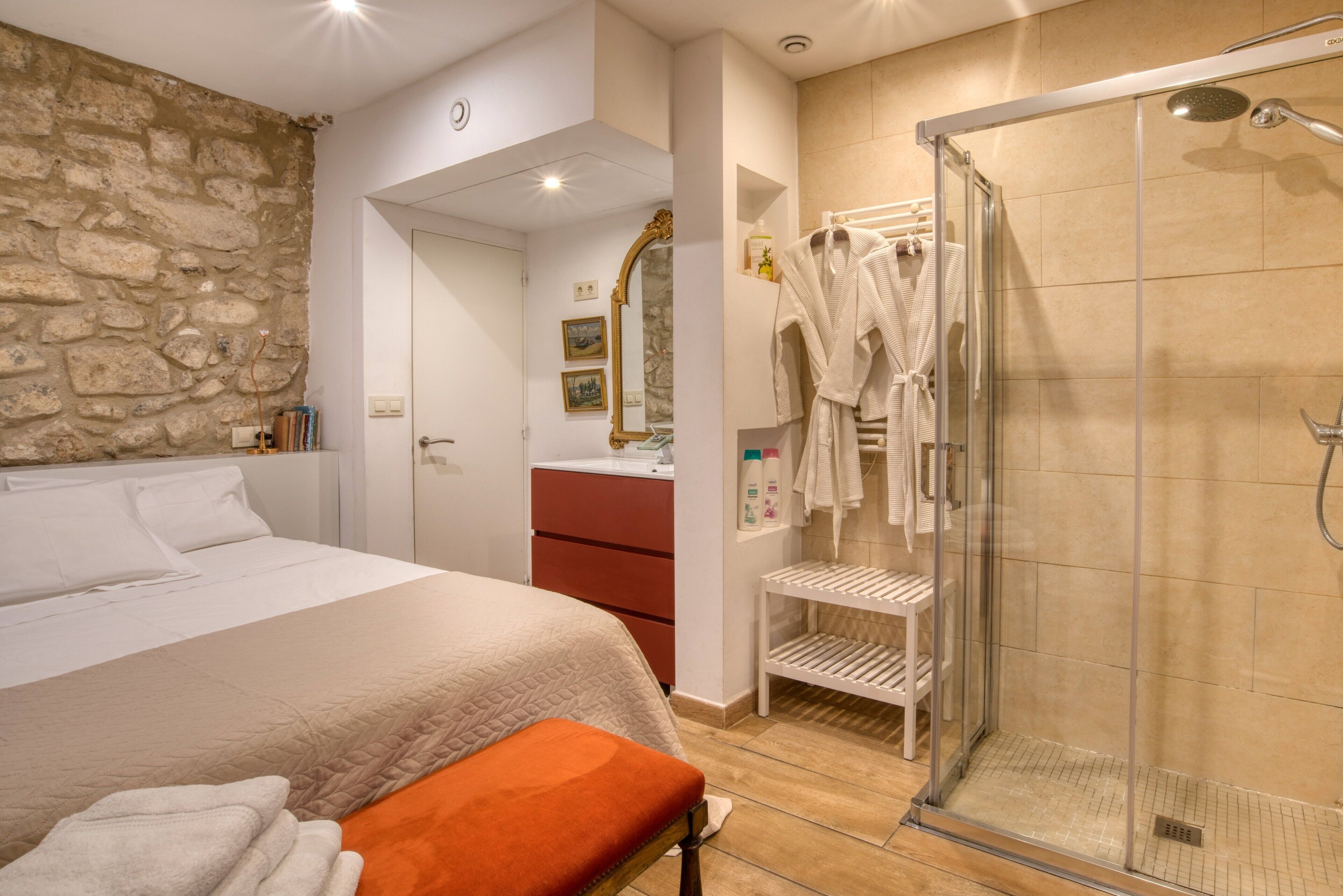 A hotel room with a bed, a shower, a bathroom with a mirror, and a stone wall.