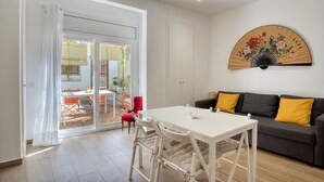Ein Zimmer mit einem weißen Tisch, einem grauen Sofa mit gelben Kissen, einem ventilator mit floralem Muster und einer Schiebetür zu einem Außenbereich.