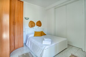 Une chambre à coucher avec un lit, du linge de lit blanc, une armoire en bois et une petite étagère fixée au mur.