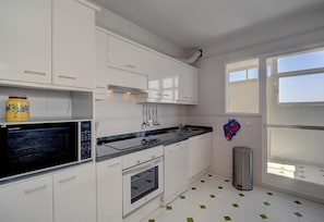 Une cuisine moderne avec des meubles blancs, un plan de travail noir, un four à micro-ondes, un four traditionnel et une fenêtre avec des stores.